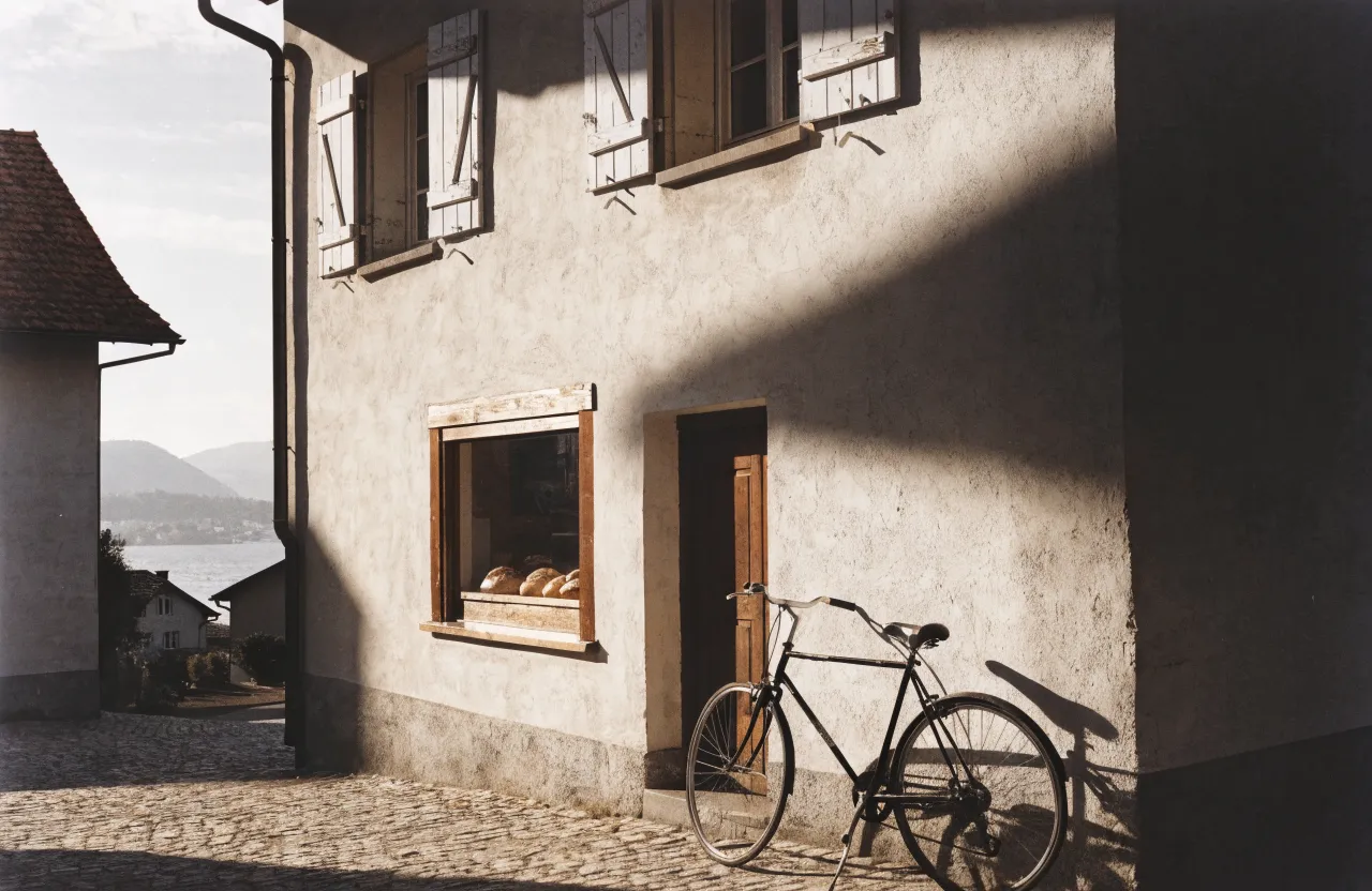 Ladenfront einer alten Dorfbäckerei mit Holzfenster voller Brot, Holztür, Kopfsteinpflaster und einem Velo an der Wand, dahinter der See im Morgenlicht.